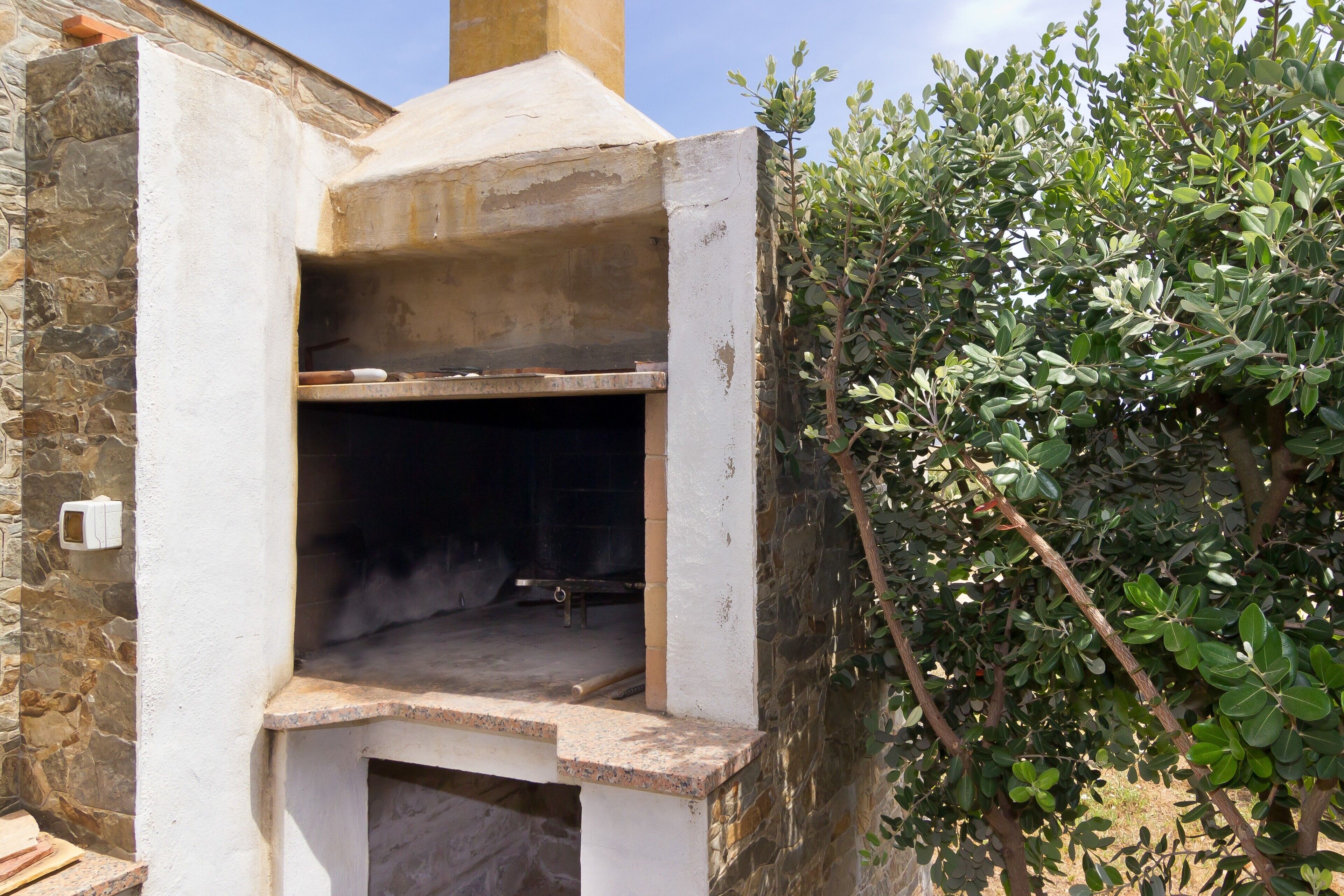 An outdoor stone oven structure with a chimney, surrounded by stone walls and a bush.