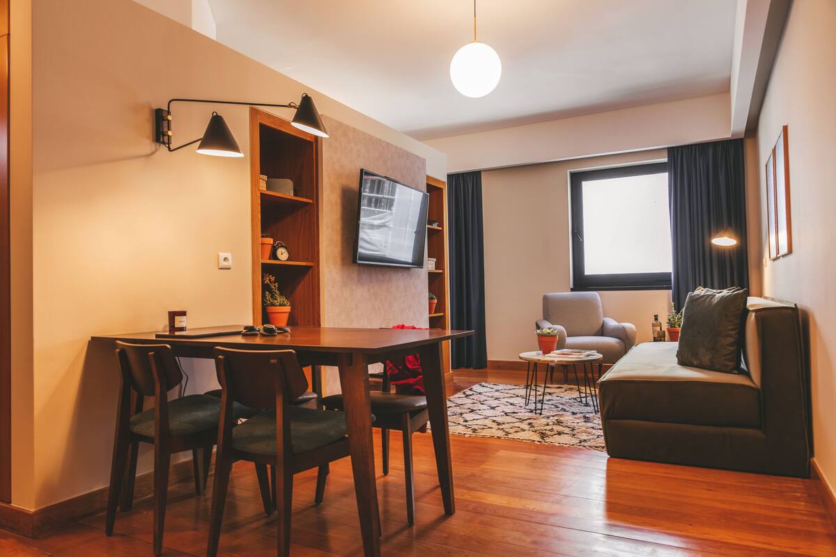 Un salon moderne avec une table à manger en bois, deux chaises, un canapé, un téléviseur fixé au mur et une grande fenêtre avec des rideaux.