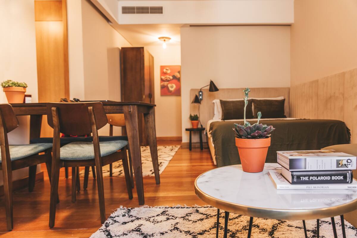 Un salon chaleureux avec une table à manger en bois, des chaises, une table basse ronde sur laquelle sont posés des livres, une plante verte et un lit avec une tête de lit en bois.