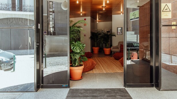 Un hall d’hôtel moderne avec une entrée en verre, des plantes en pot, un plafond en bois et un escalier apparent.