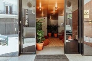 Um hall de hotel moderno com entrada de vidro, plantas em vasos, teto de madeira e escadaria visível.