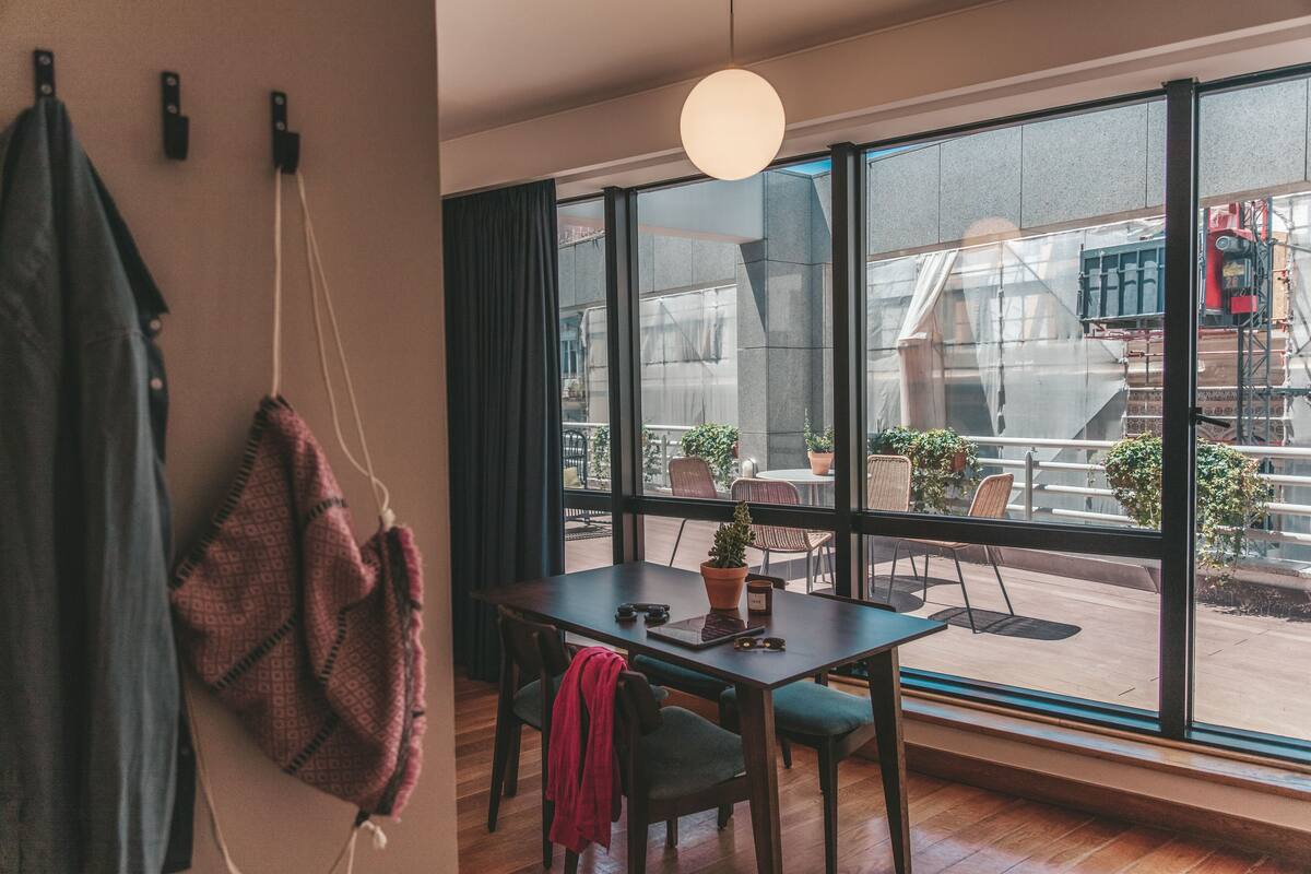 Une chambre moderne dotée d’une grande fenêtre, d’un plancher en bois, d’une table avec des chaises et d’un porte-manteau suspendu.