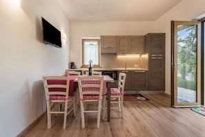 A compact kitchen with wooden cabinets, a dining table with chairs, a flat-screen TV mounted on the wall, and a sliding glass door leading to an outdoor area.
