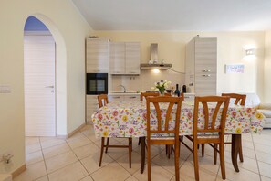 A dining area with a table set for four, surrounded by wooden chairs, in a room with a tiled floor and a kitchen in the background. - Holiday Apartment "Elga" with Garden & Wi-Fi (Villasimius)