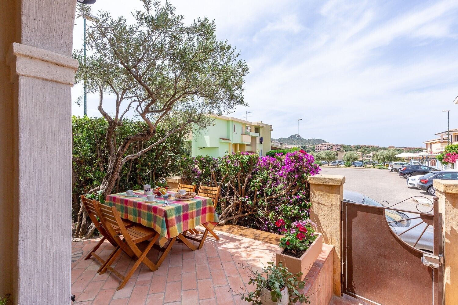 An outdoor patio with a table and chairs, surrounded by plants and a view of a parking area and buildings.