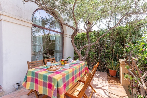 An outdoor dining area with a table set for breakfast, surrounded by greenery and a tree.