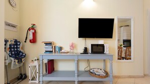 Una habitación con una televisión montada en la pared, una mesita con libros y un microondas, un extintor, y una silla con funda estampada.