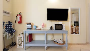 A room with a television mounted on the wall, a small table with books and a microwave, a fire extinguisher, and a chair with a patterned cover.
