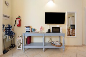 A room with a television mounted on the wall, a small table with books and a microwave, a fire extinguisher, and a chair with a patterned cover. - Holiday Apartment "Elga" with Garden & Wi-Fi (Villasimius)