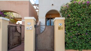 Una entrada protegida con un portón metálico y una puerta de madera, flanqueada por dos letreros numerados, que conduce a un edificio con fachada color crema y un seto verde.