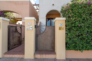 A gated entrance with a metal gate and a wooden door, flanked by two numbered signs, leading to a building with a beige facade and a green hedge. - Holiday Apartment "Elga" with Garden & Wi-Fi (Villasimius)