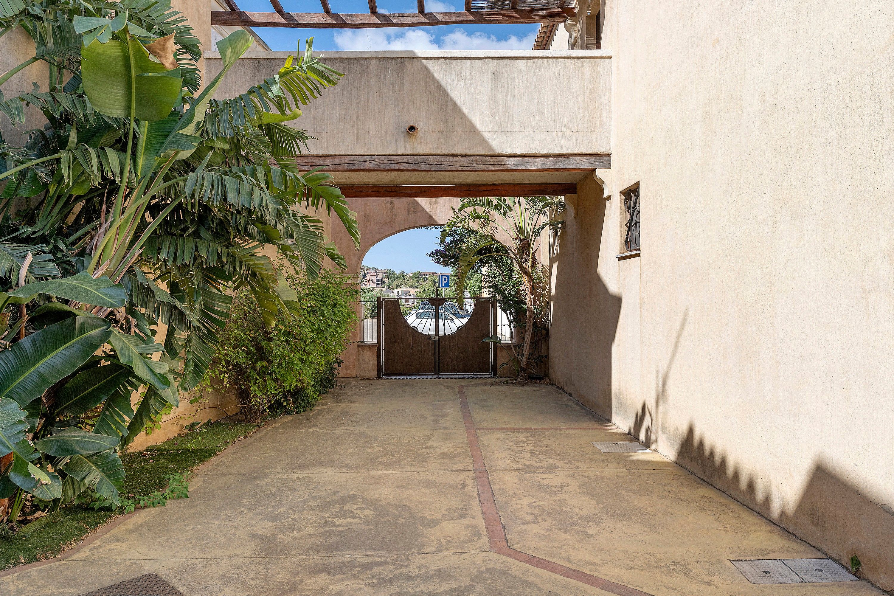 A paved pathway leading to a gated entrance with a circular window, surrounded by lush greenery and a building with a flat facade.