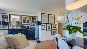 Ein Wohnzimmer mit einer Couch, einem Esstisch, Stühlen, einer großen Vase mit Blumen, einer Bar mit Flaschen, gerahmten Bildern an der Wand und einer Küchenzeile.