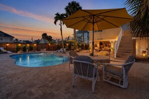 A swimming pool with lounge chairs and a table, surrounded by a wooden fence and palm trees.