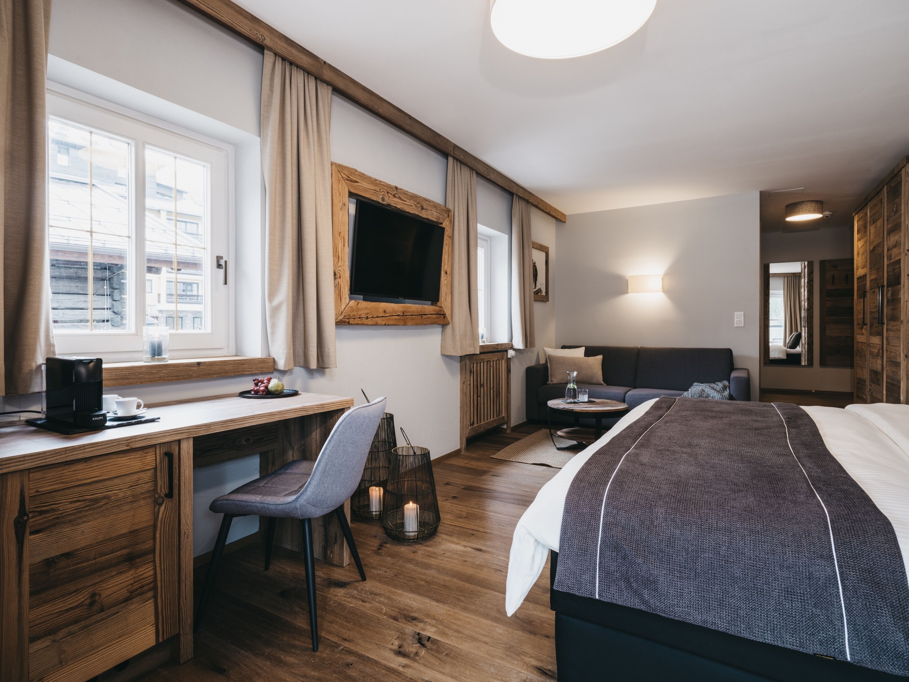 Een moderne hotelkamer met een houten bureau, een comfortabele stoel, een bed met een donker hoofdbord, een nachtkastje met een lamp en een groot raam met gordijnen.