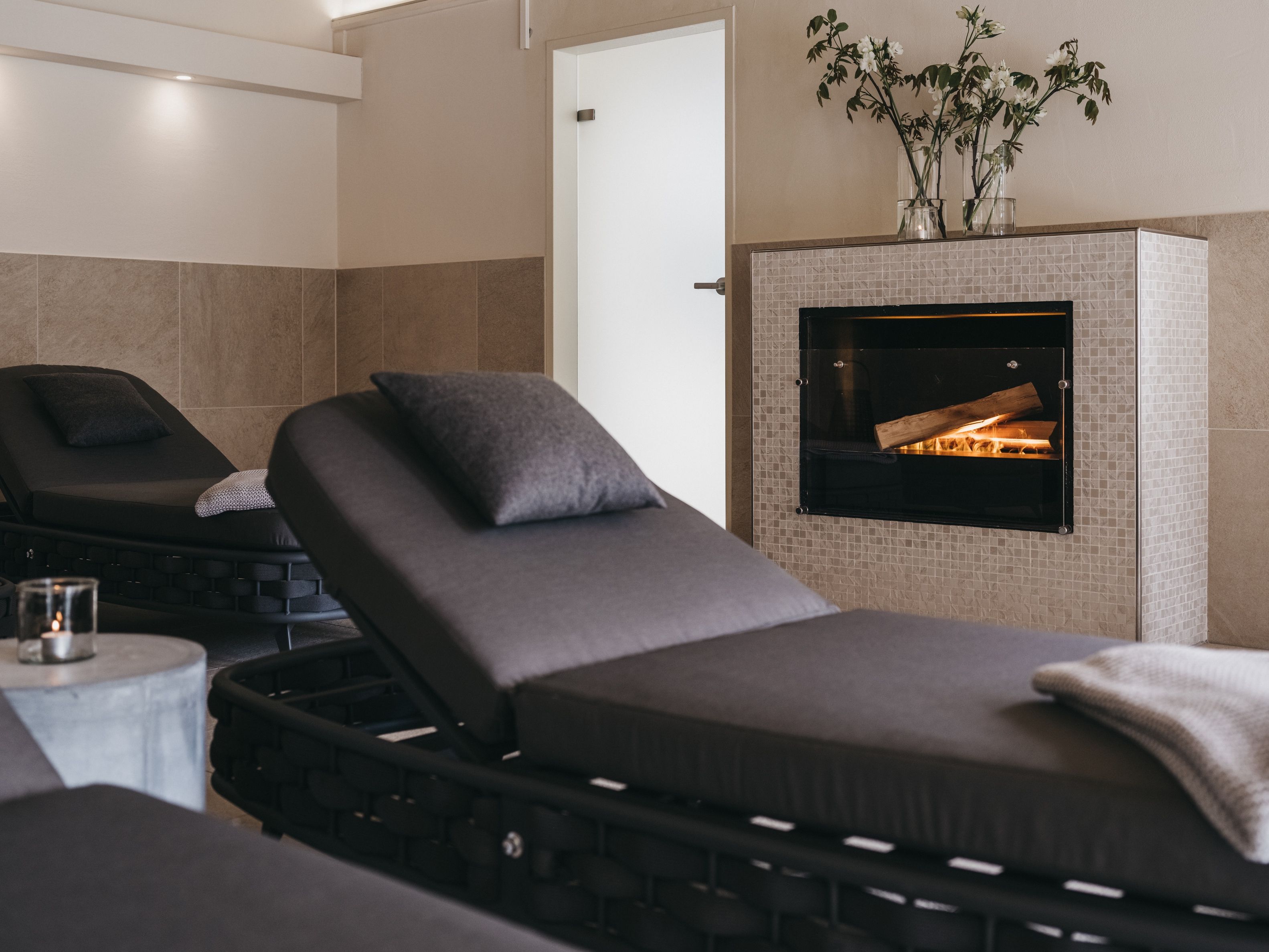 A spa room with two massage chairs, a small table with a glass, and a fireplace with a plant on top.