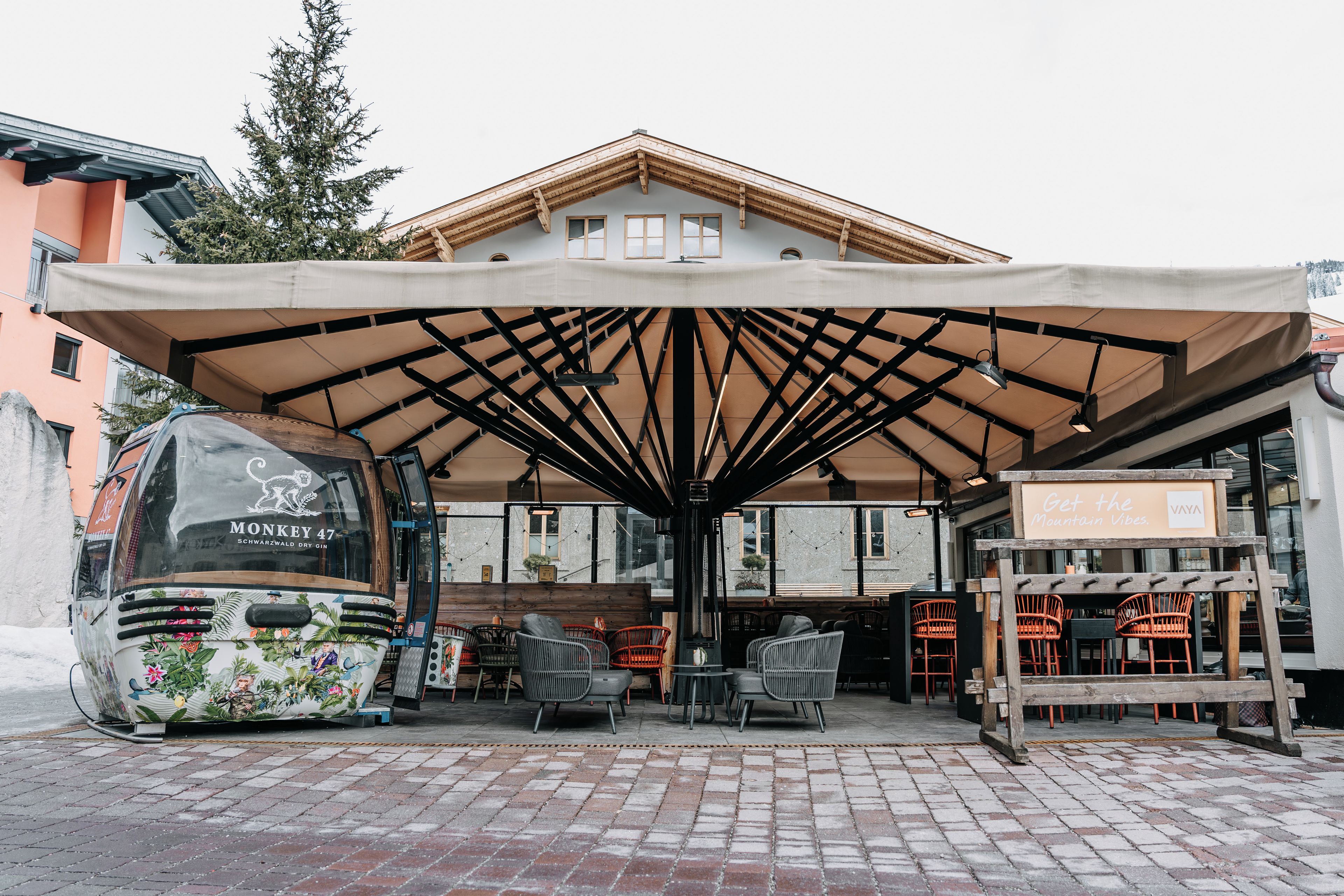 A covered outdoor seating area with tables and chairs, a bus parked to the side, and a building with a sloped roof in the background.