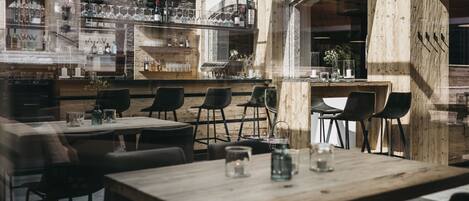A restaurant interior with wooden tables and chairs, a bar area with bottles and glasses, and a view through a window.