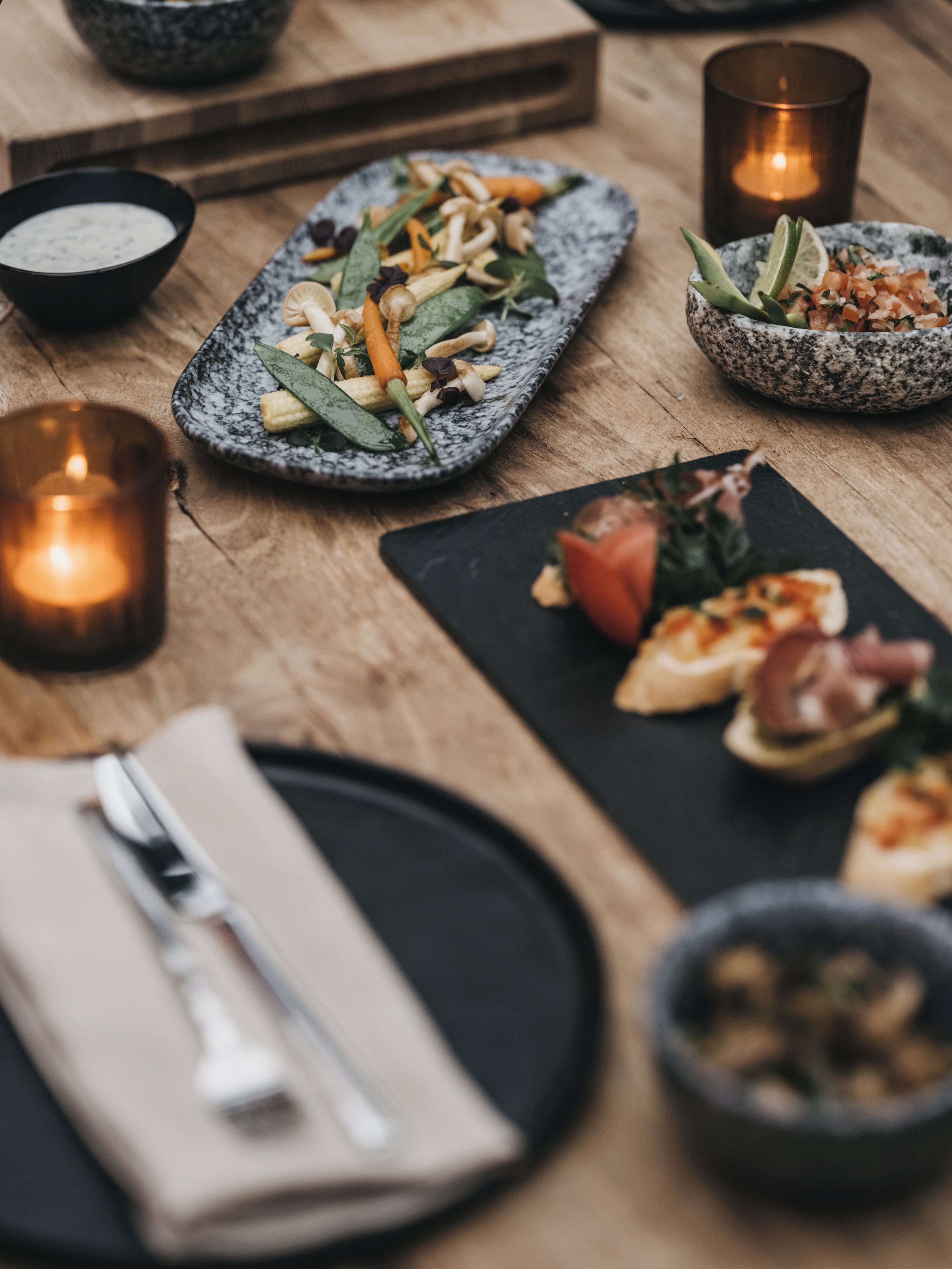 Ein Tisch ist reichhaltig gedeckt mit verschiedenen Speisen, darunter ein Salat, eine Suppenschüssel und eine Platte mit Vorspeisen. Kerzen und eine Oliven-Schale stehen auf dem Tisch.