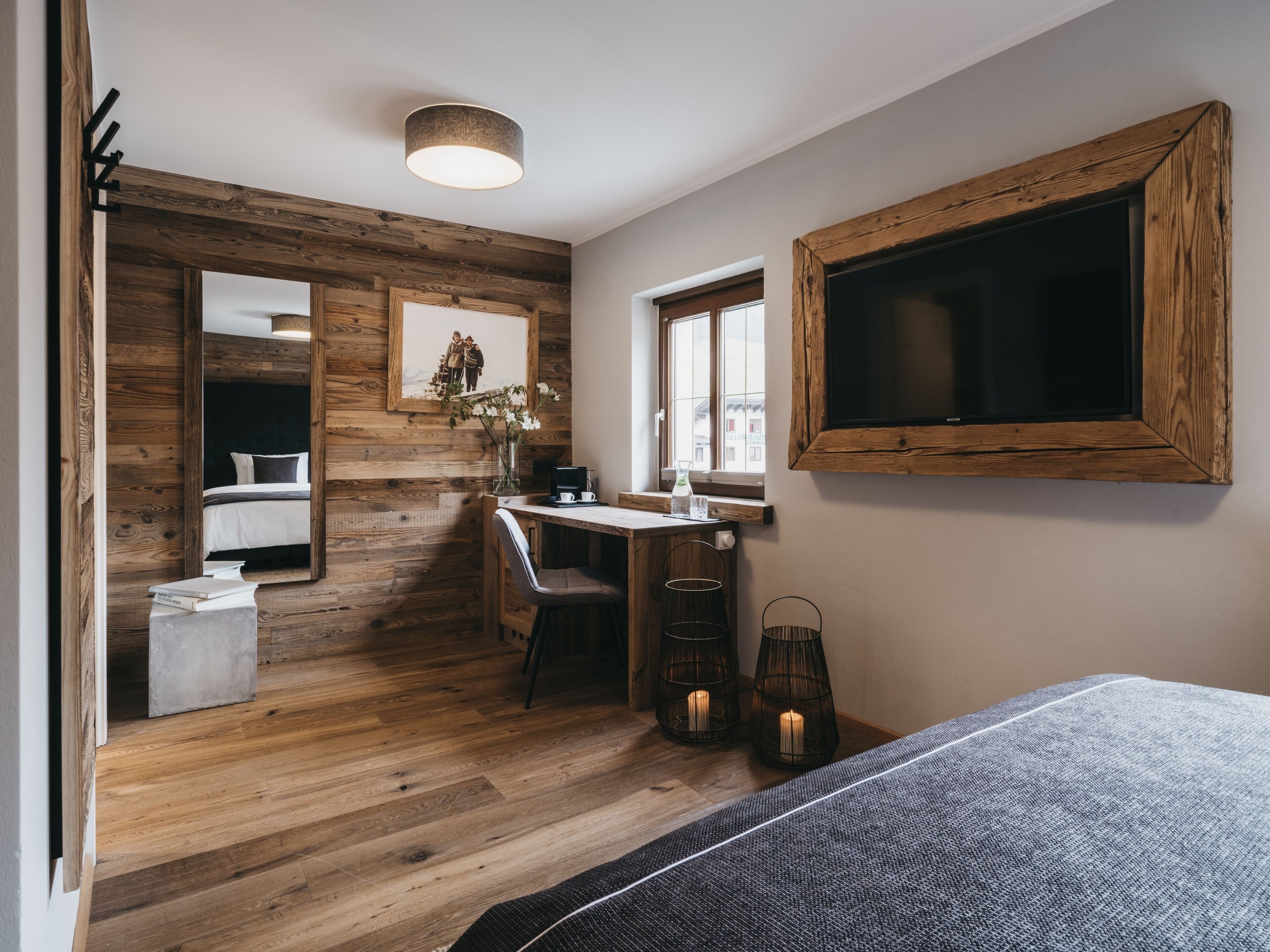A room with a wooden floor, a bed, a desk with a chair, a large mirror, a window, and a wall-mounted TV.