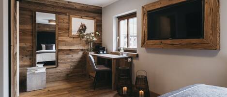 Une pièce avec un plancher en bois, un lit, un bureau avec une chaise, un grand miroir, une fenêtre et un téléviseur fixé au mur.