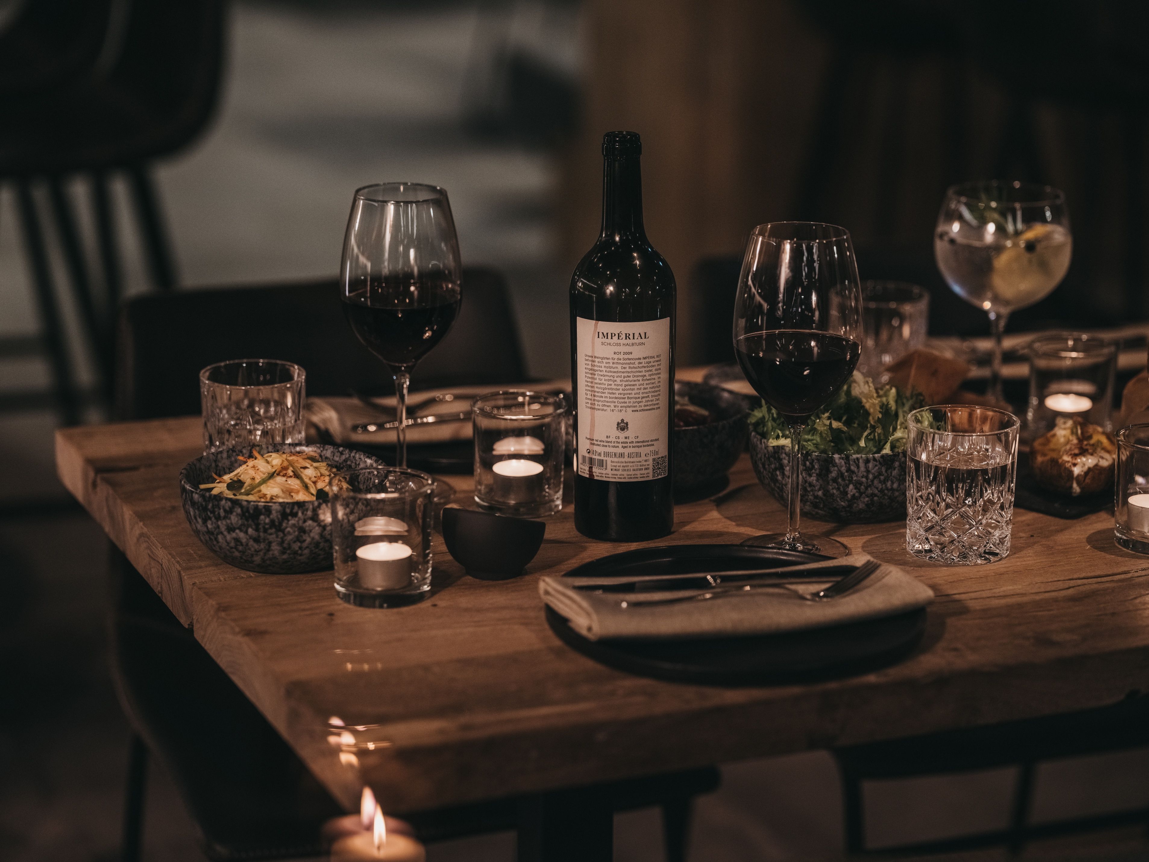 A wooden table set with wine glasses, a bottle, candles, and a bowl of food.