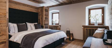 Un dormitorio con techo de madera, una cama grande con cabecera oscura, un banco de madera y dos ventanas adornadas con arreglos florales.