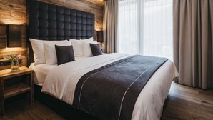 Quarto moderno com cabeceira de madeira, uma cama grande com roupa de cama branca e escura, mesa de cabeceira com um vaso e uma janela ampla com cortinas.
