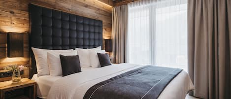 A modern bedroom with a wooden headboard, a large bed with white and dark bedding, a bedside table with a vase, and a large window with curtains.