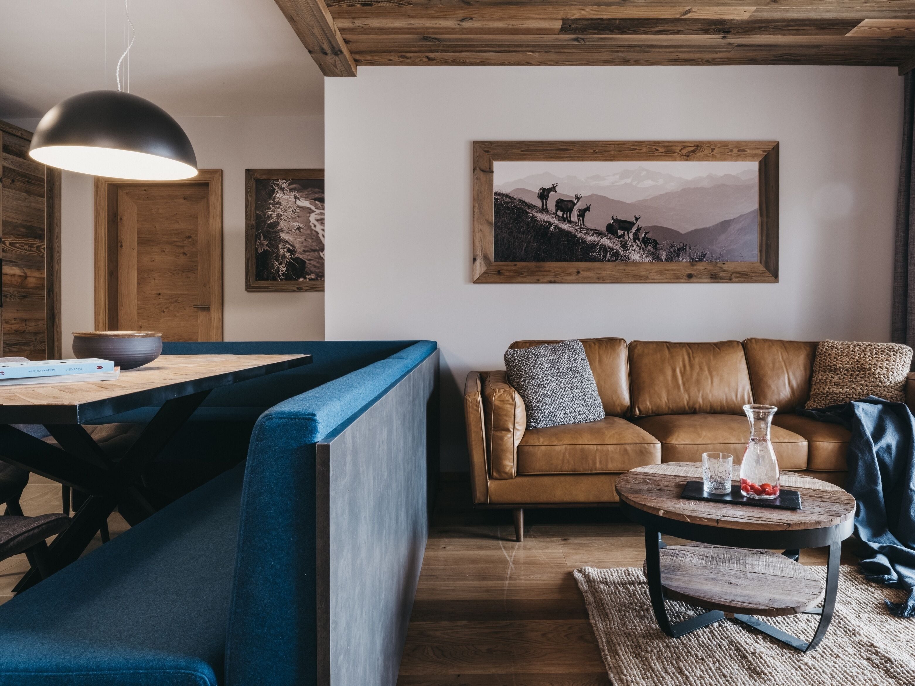 A cozy living area with a wooden ceiling, a brown leather sofa, a blue bench, a wooden coffee table, and a painting of deer on a mountain.