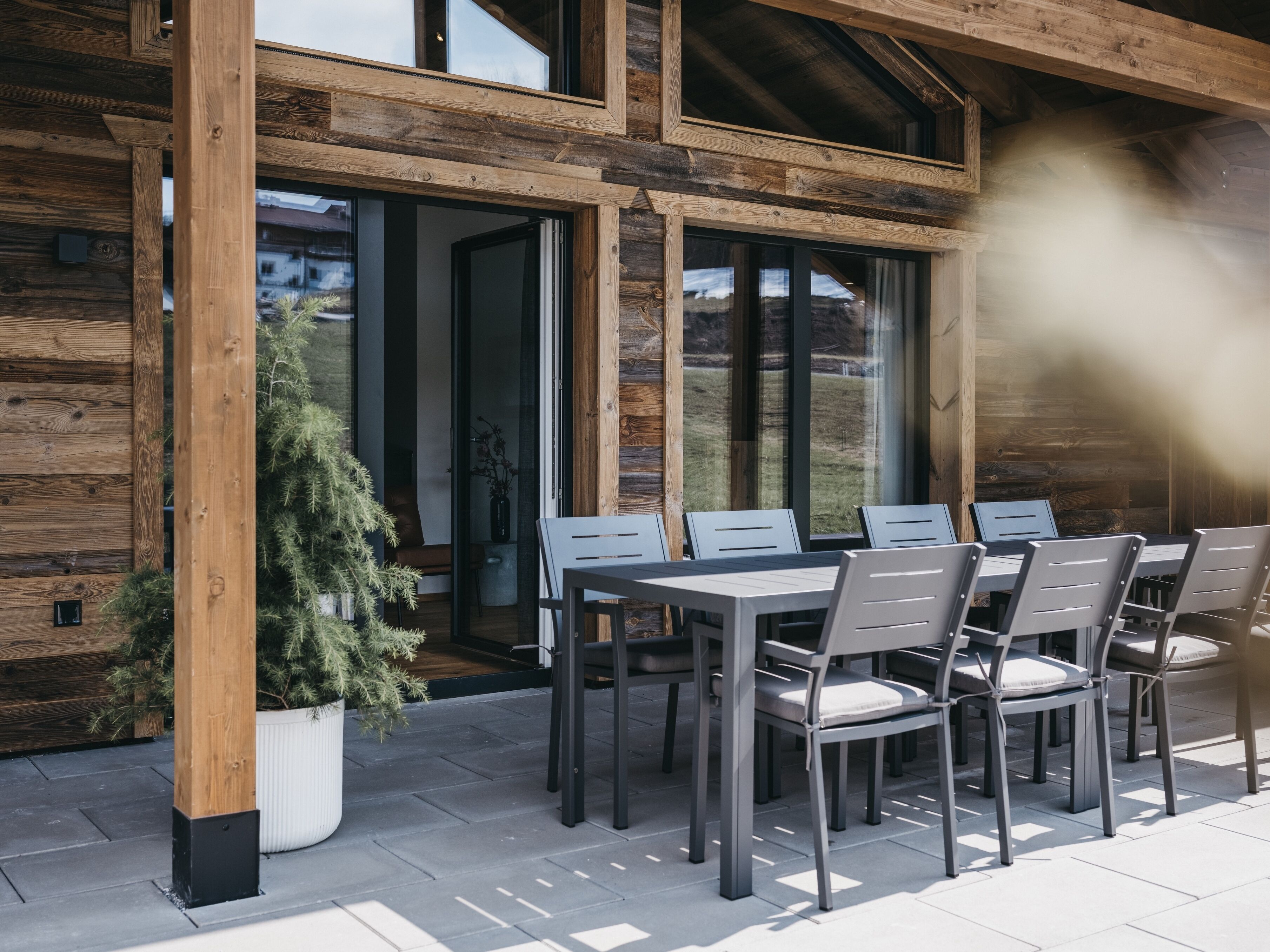 An outdoor patio with a wooden table and chairs, a potted plant, and a view of a residential area.