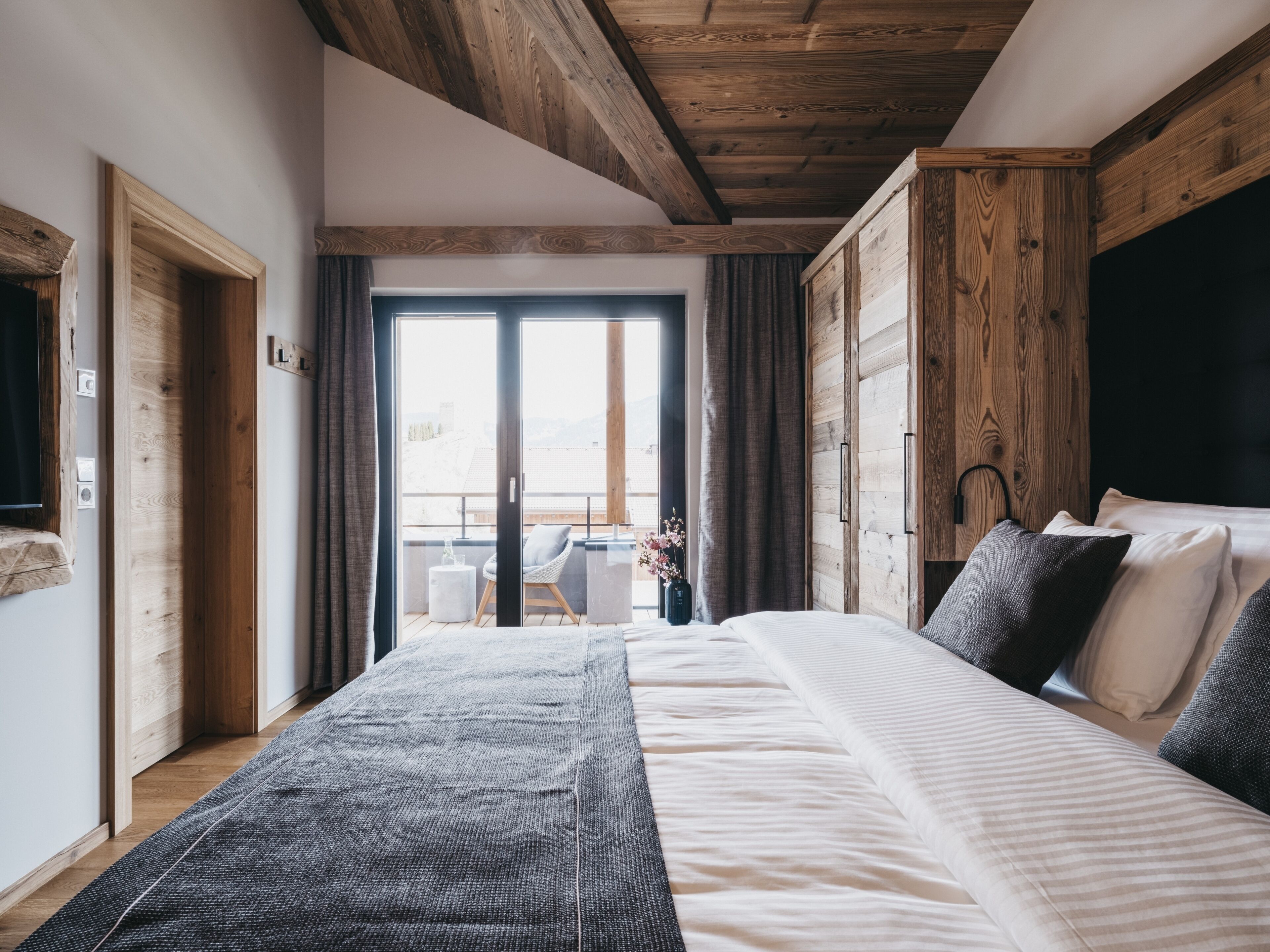 Une chambre avec un grand lit, des poutres apparentes en bois au plafond, une vue à travers une porte coulissante et une petite table avec une chaise à l’extérieur.