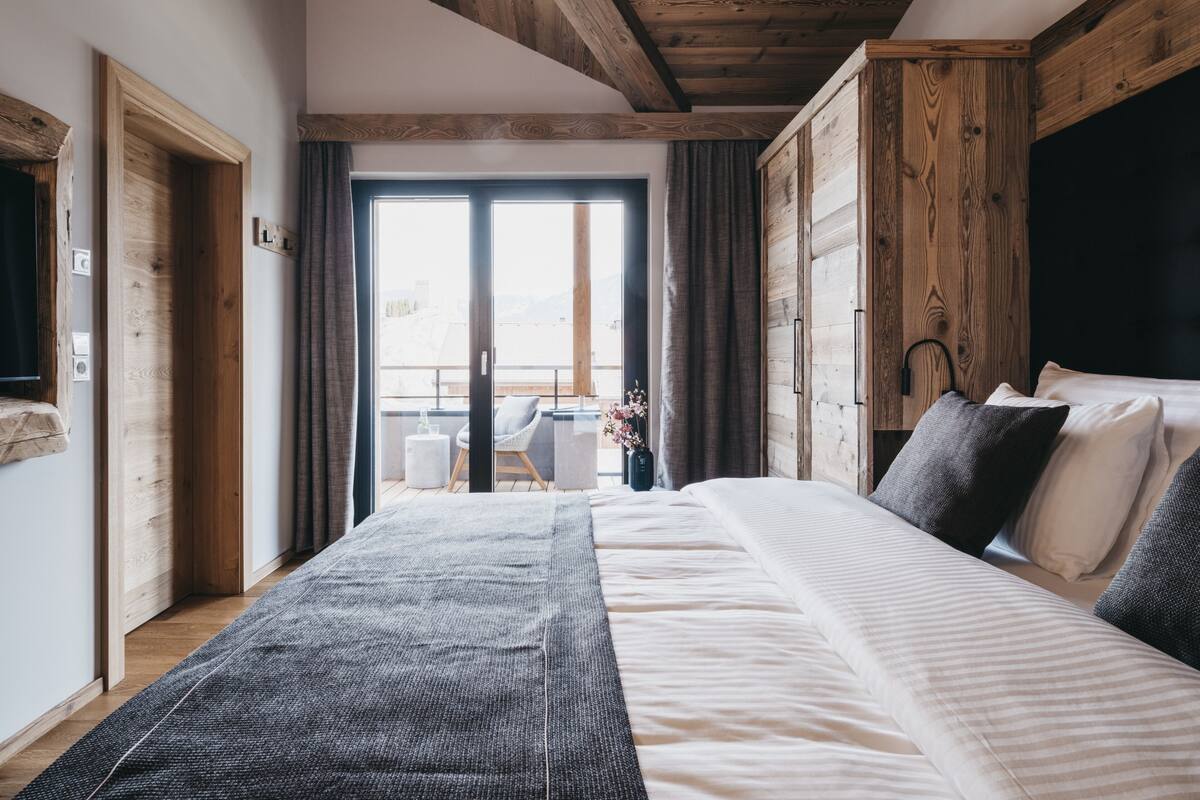 A bedroom with a large bed, wooden ceiling beams, a view through a sliding door, and a small table with a chair outside.