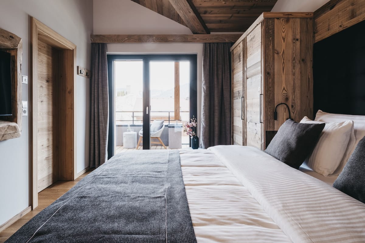 Un dormitorio con una cama grande, vigas de madera en el techo, vista a través de un puertas corredera, y una pequeña mesa con una silla afuera.