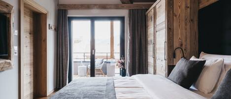 Ein Schlafzimmer mit einem großen Bett, Holzbalkendecke, Blick durch eine Schiebetür und einem kleinen Tisch mit Stuhl davor.