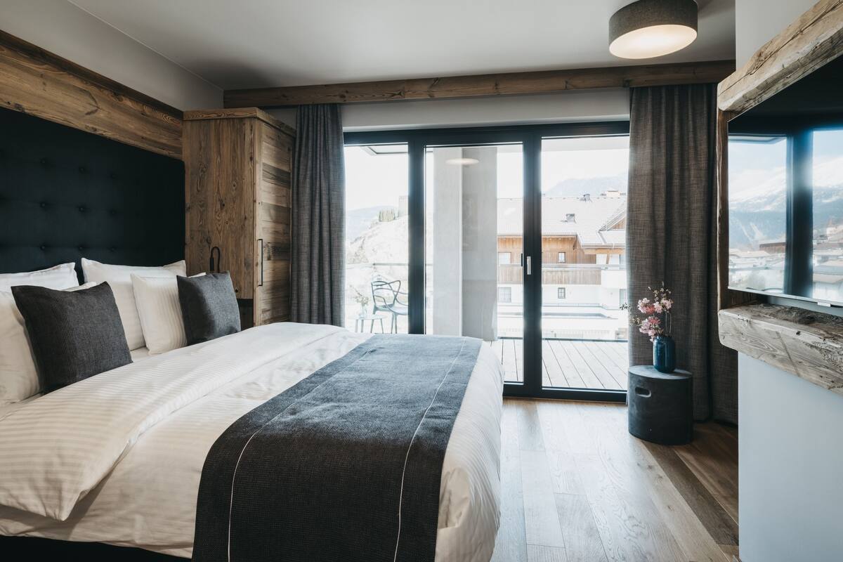 A modern bedroom with a large bed, wooden headboard, and a view of a balcony with a cityscape.