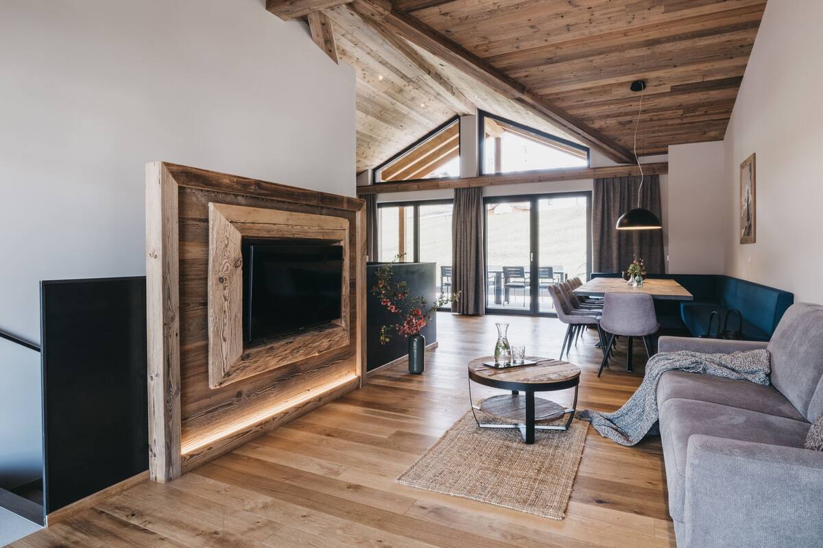 A modern living room with a wooden ceiling, a large TV mounted on a wooden frame, a round coffee table, and a sofa.