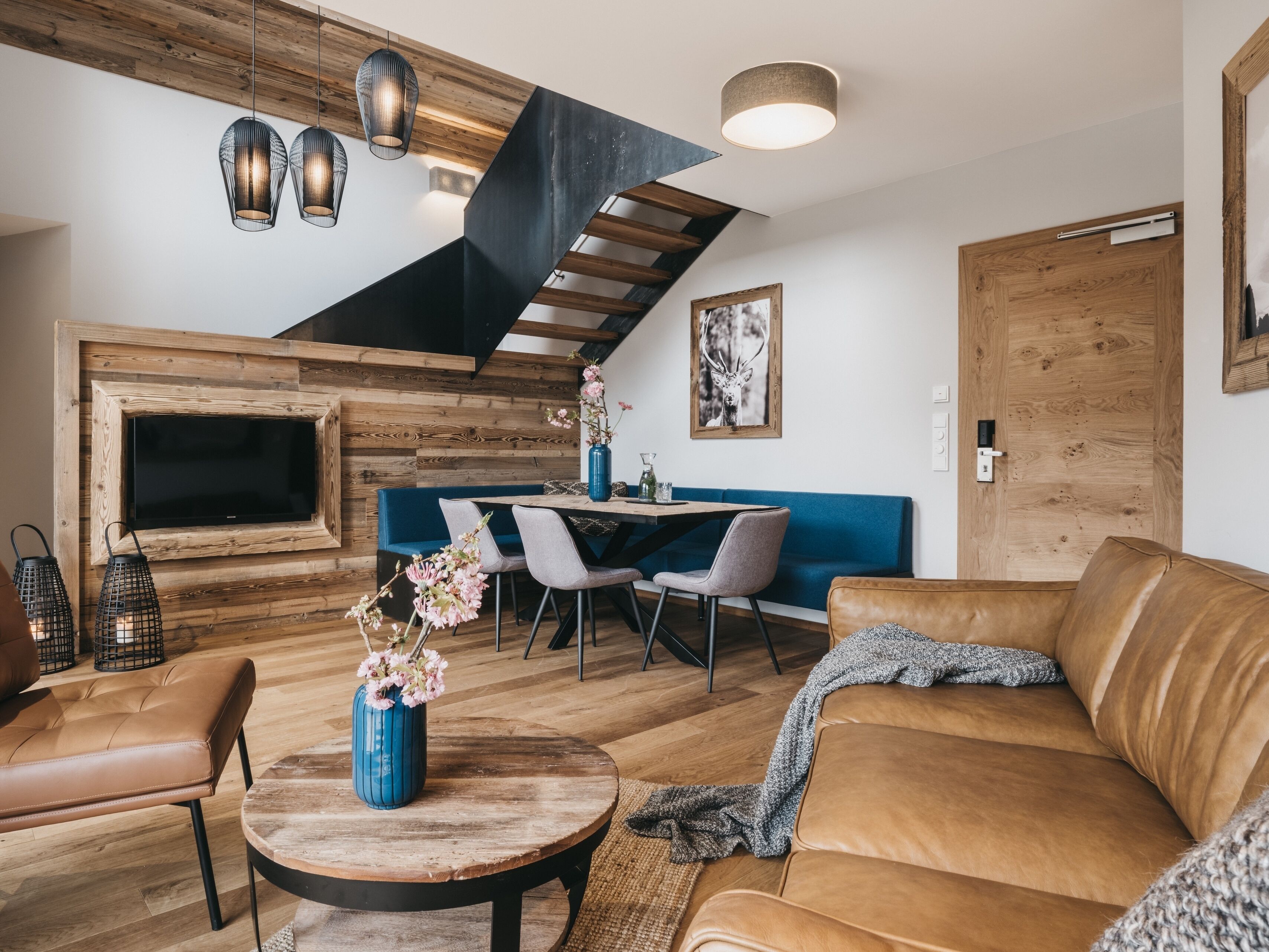A modern living room with a wooden accent wall, a blue sofa, a round coffee table with a vase of flowers, and a dining area with a blue bench and chairs.