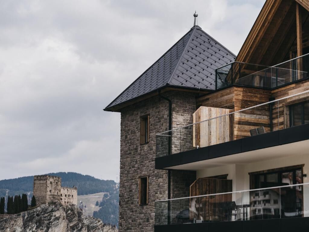 A modern building with a stone facade and a wooden balcony, overlooking a historic castle and a mountainous landscape.