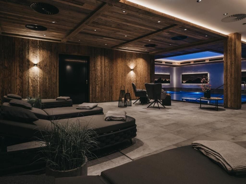 Une salle de spa moderne avec un plafond en bois, une table de massage, un fauteuil de massage et un espace d’accueil doté d’un mur bleu et donnant sur la piscine.