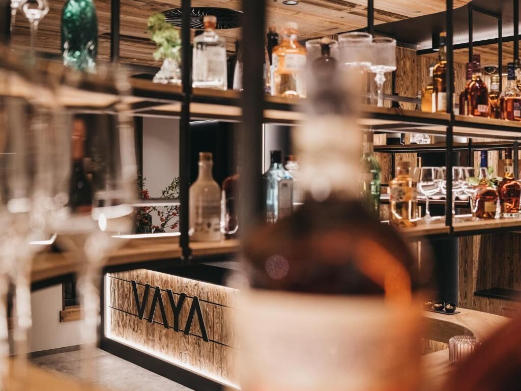A bar area with shelves of bottles and glasses, a wooden counter, and a sign with the letters "VAY".