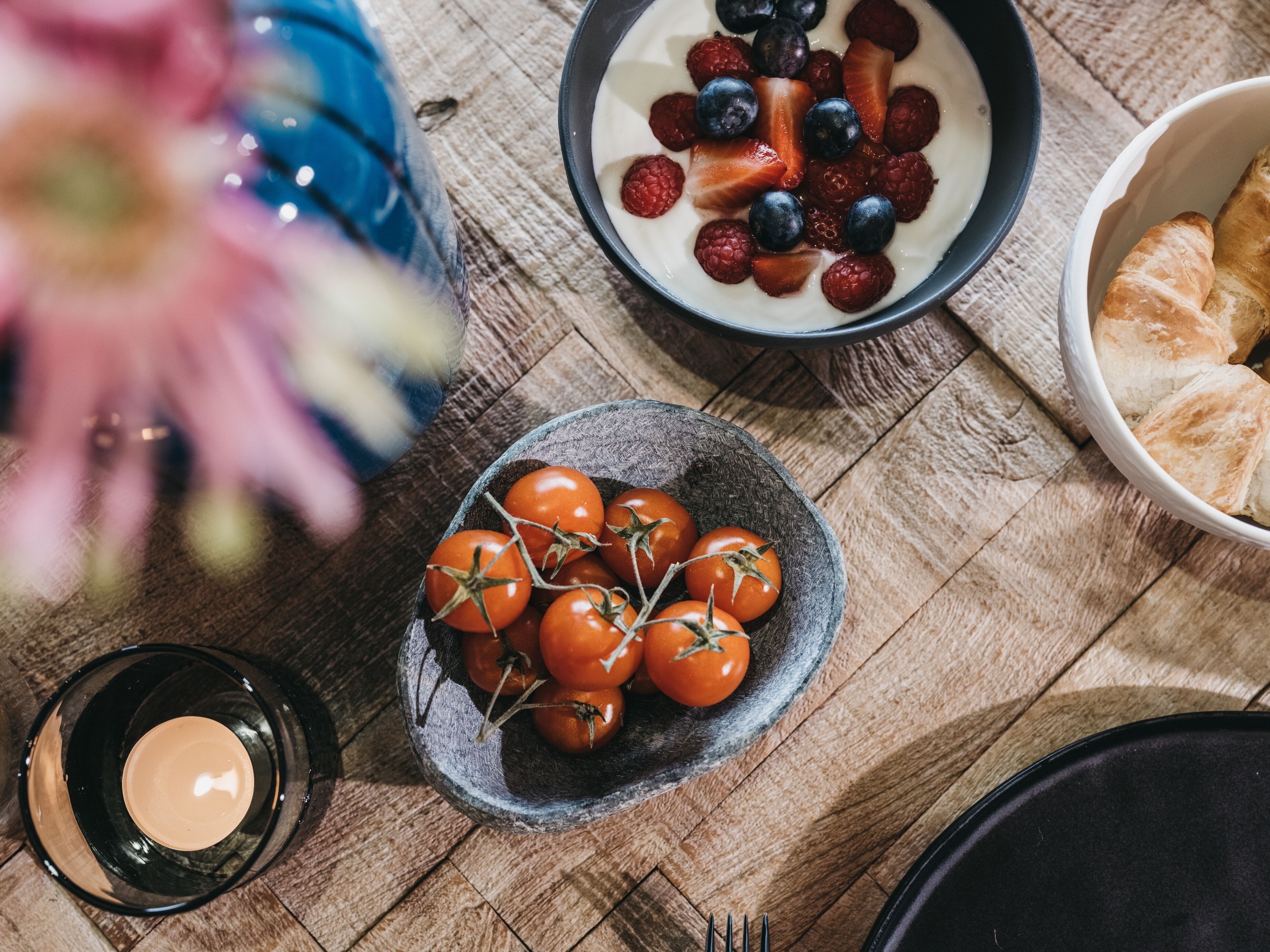 A wooden table with a bowl of cherry tomatoes, a plate of berries, a candle, and a bowl of croissants.