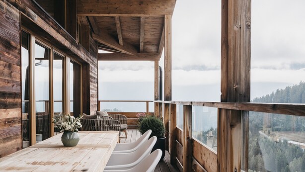Een houten terras met een tafel en stoelen, grote ramen en uitzicht op een bebost dal.