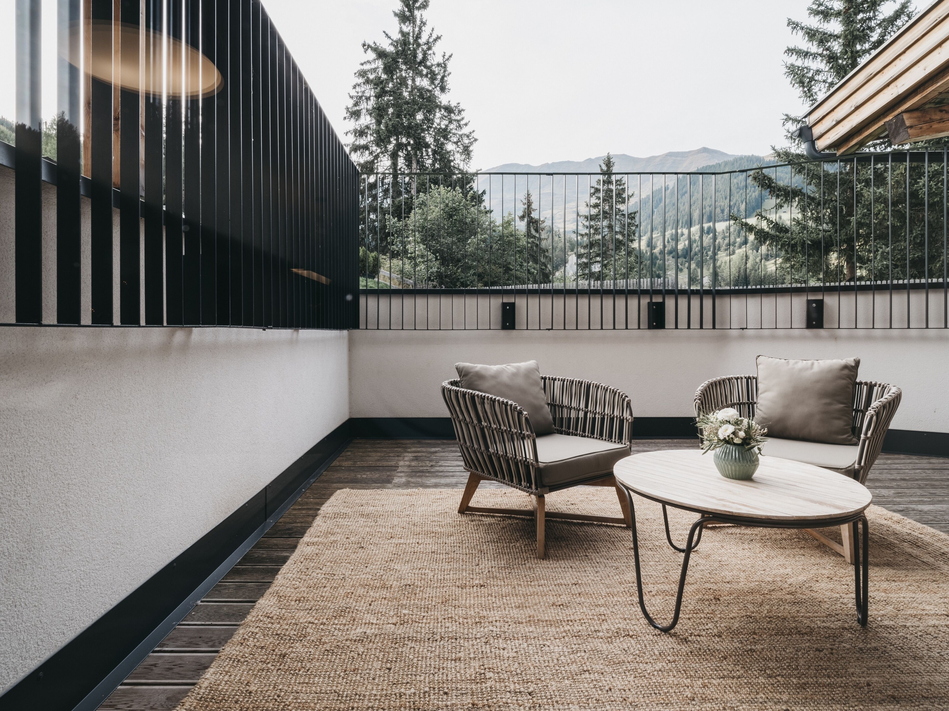 A modern outdoor seating area with two wicker chairs and a round table, overlooking a mountainous landscape.