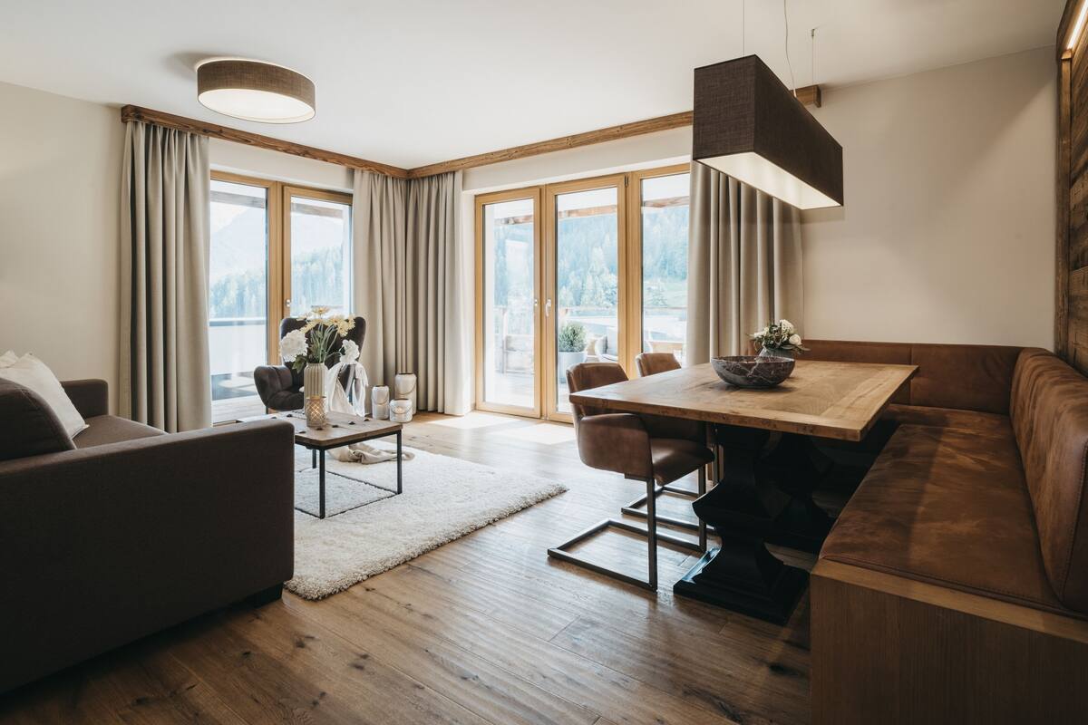 A modern living room with a wooden floor, a brown leather sofa, a dining table with chairs, and large windows with curtains.