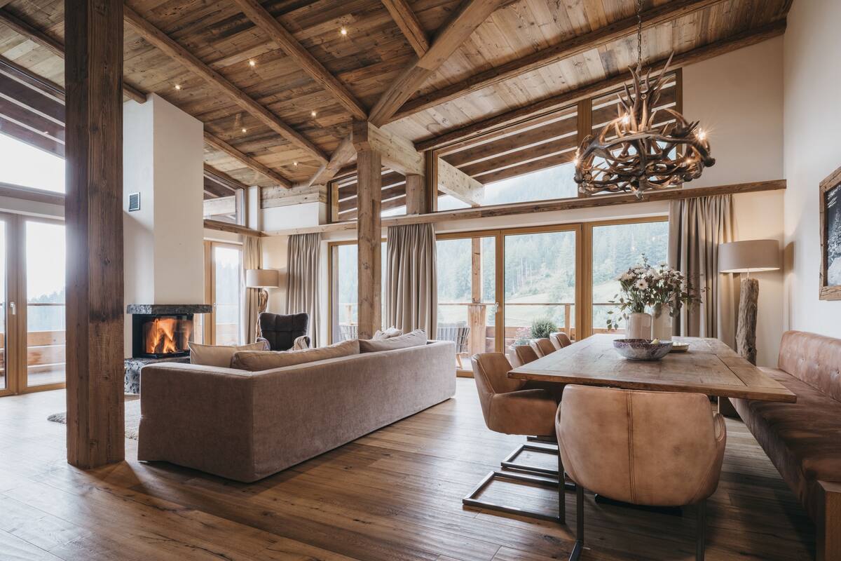 A spacious living area with a fireplace, wooden beams, a dining table, and large windows offering a view of the outdoors.