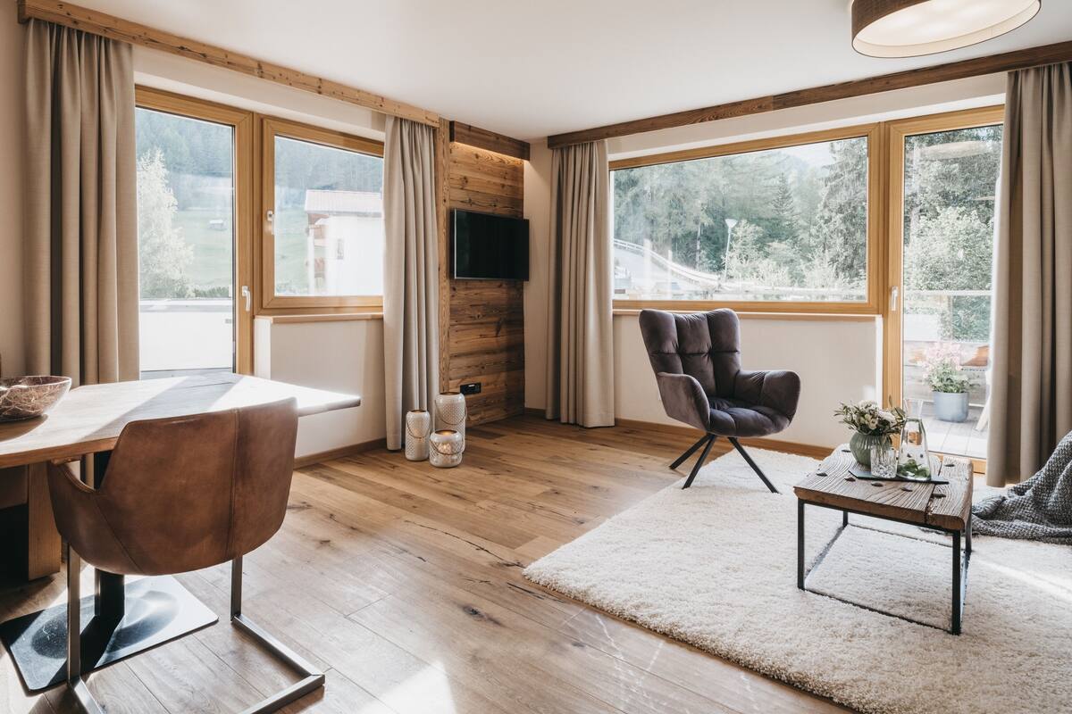 A modern living room with wooden flooring, a brown leather chair, a white rug, a wooden coffee table, and large windows with curtains.