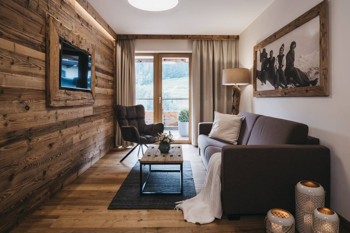 A cozy living room with a wooden wall, a sofa, a chair, a coffee table with flowers, and a painting on the wall.
