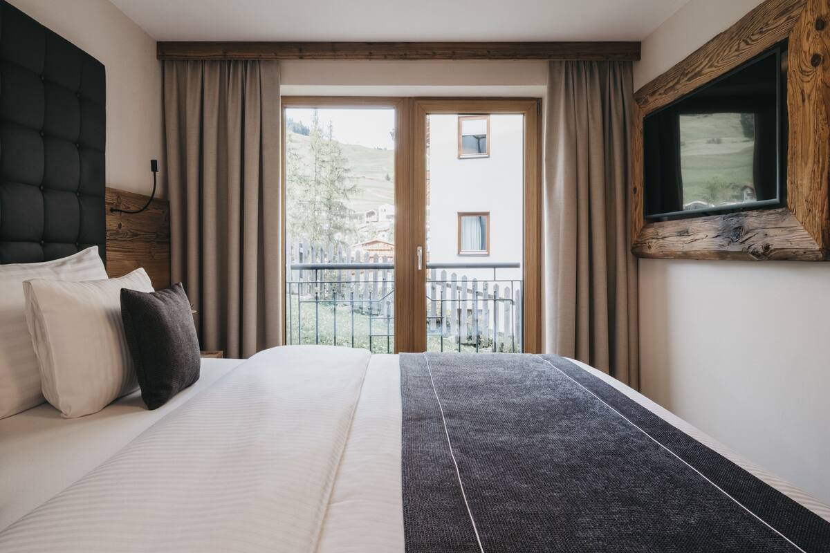 A bedroom with a large bed, a wooden headboard, a TV mounted on the wall, and a view of a balcony with a fence.