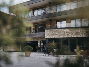 A modern multi-story building with a stone facade, large windows, and outdoor seating area. - VAYA St. Anton (Sankt Anton am Arlberg)